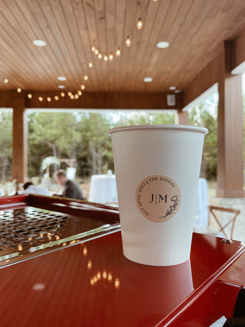 A cup of coffee placed on a red espresso machine with white table arranged for a wedding coffee cart, highlighting mobile coffee bar rental options of personalized stickers on cups in Waycross, Georgia.