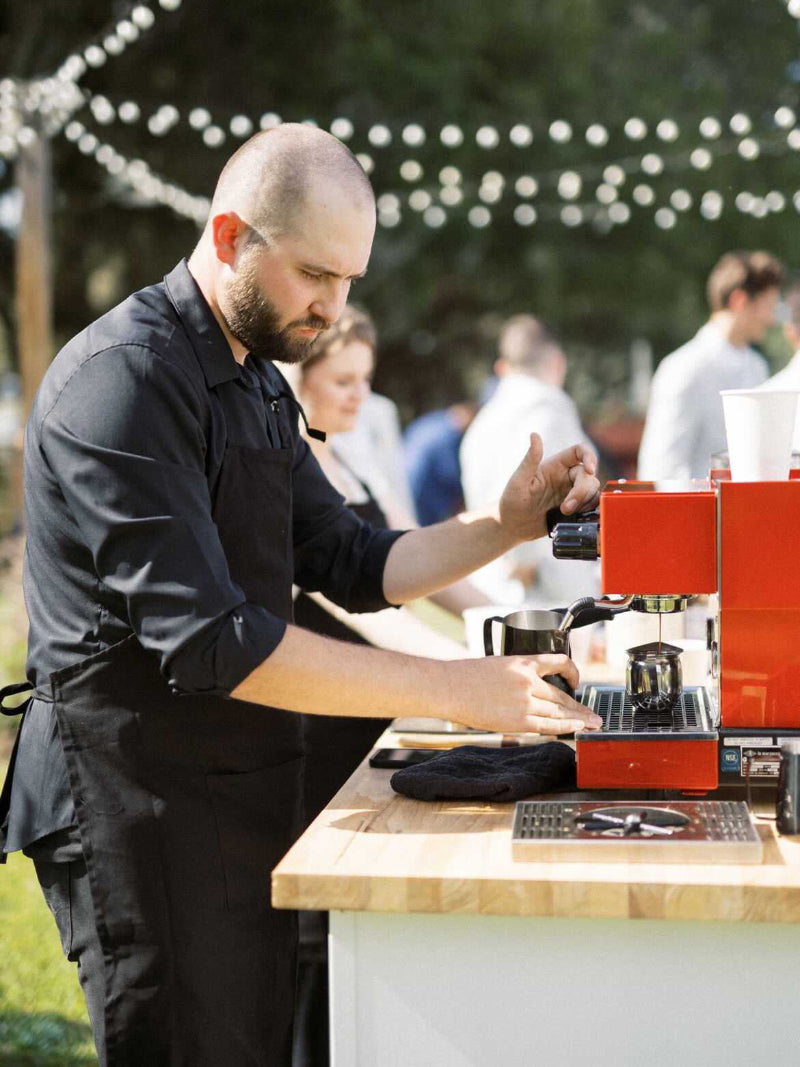 A barista in a black apron operates a red espresso machine at a mobile espresso bar, serving guests at an outdoor coffee station adorned with string lights.